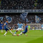 Jogadores de Chelsea e Manchester City disputando a bola em um jogo de futebol, com torcedores ao fundo.